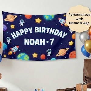 May include: A navy blue birthday banner with a space theme, featuring planets, rockets, astronauts, and stars. The banner reads "HAPPY BIRTHDAY NOAH • 7". Balloons in silver, orange, gold, and blue are on the left and right sides of the banner.