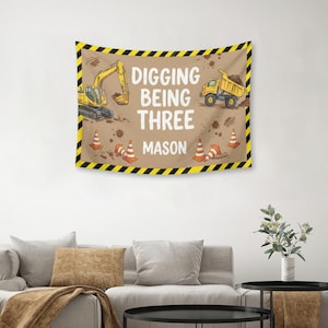 Puede incluir: Un tapiz beige con tem&aacute;tica de construcci&oacute;n. Presenta una excavadora y un cami&oacute;n volquete amarillos, conos de tr&aacute;fico naranjas y el texto "DIGGING BEING THREE MASON" en blanco. El borde es negro y amarillo.