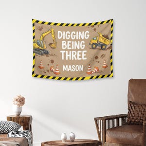 Puede incluir: Un tapiz marr&oacute;n con tem&aacute;tica de construcci&oacute;n. Presenta una excavadora y un cami&oacute;n volquete amarillos, conos de tr&aacute;fico naranjas y el texto "DIGGING BEING THREE MASON" en blanco. El borde es negro y amarillo.
