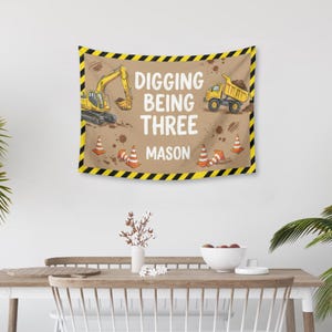 Puede incluir: Un tapiz marr&oacute;n con un tema de construcci&oacute;n, con una excavadora y un cami&oacute;n volquete amarillos. El texto "Digging Being Three Mason" se muestra en blanco. El borde es de rayas negras y amarillas, con conos de tr&aacute;fico naranjas.