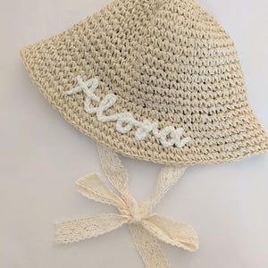 May include: A beige straw sun hat with a white lace ribbon tied in a bow. The hat has the word "Aloha" embroidered on the front in white thread.