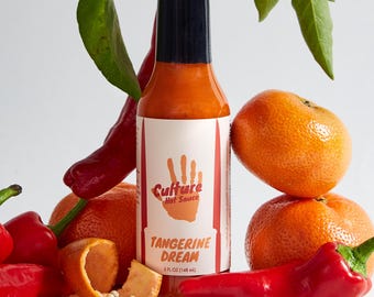 Op de afbeelding: Een fles Culture Hot Sauce met het label "Tangerine Dream" en een handafdruklogo. De fles is omringd door rode pepers, witte peperkorrels en oranje mandarijnen.