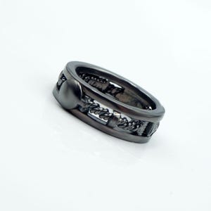Puede incluir: Un anillo gris oscuro con un diseño texturizado. El anillo presenta una serie de formas interconectadas y un acento redondeado. El anillo está sobre un fondo blanco.