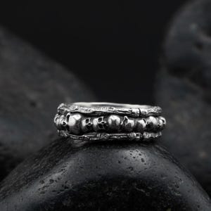 Op de afbeelding: Een zilveren ring met een herhaald schedelontwerp. De ring heeft een gedetailleerd, getextureerd uiterlijk, met meerdere kleine schedels die de band omcirkelen. De ring wordt getoond op een donkere, getextureerde ondergrond.