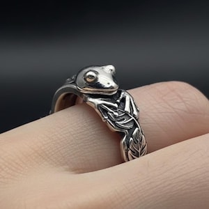 May include: A silver ring with a frog design. The frog sits on the ring's band, with detailed leaf patterns. The ring is displayed on a finger against a dark backdrop.
