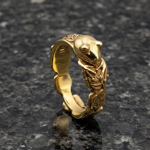 Op de afbeelding: Een goudkleurige ring met een kikkerontwerp. De ring heeft een gedetailleerde kikkerfiguur aan de bovenkant, met ingewikkelde bladeren aan de zijkanten. De ring staat tegen een donkere, getextureerde achtergrond.