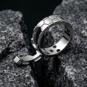 May include: A silver ring with a unique design, featuring a series of hexagonal shapes. The ring has a hinged section that opens, revealing a hidden compartment. The ring is displayed on a textured, dark gray stone surface.