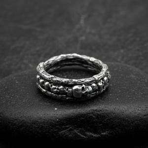 Op de afbeelding: Een zilverkleurige ring met een schedelontwerp. De ring heeft een getextureerde band en een rij kleine schedels rond het midden. De ring wordt gepresenteerd op een donker, getextureerd oppervlak.