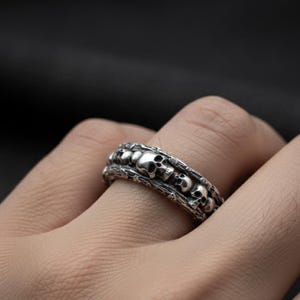 Op de afbeelding: Een zilverkleurige ring met een herhalend schedelontwerp. De ring heeft een getextureerde band en een gedetailleerd, gotisch ontwerp. De schedels zijn klein en gelijkmatig verdeeld over de ring.