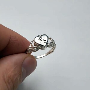 Peut inclure: Une bague argentée présentant un élément central en forme de cœur gravé des lettres AS. L'anneau est conçu avec deux mains squelettiques qui semblent tenir le cœur de chaque côté, créant une pièce en métal détaillée et structurée.