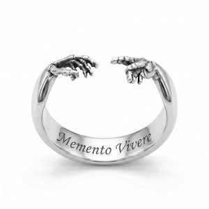 May include: A silver-toned open ring featuring a skeletal hand reaching toward a human hand. The inner band is engraved with the text Memento Vivere in a serif font. The metal has a polished finish with darkened recessed details on the hands.