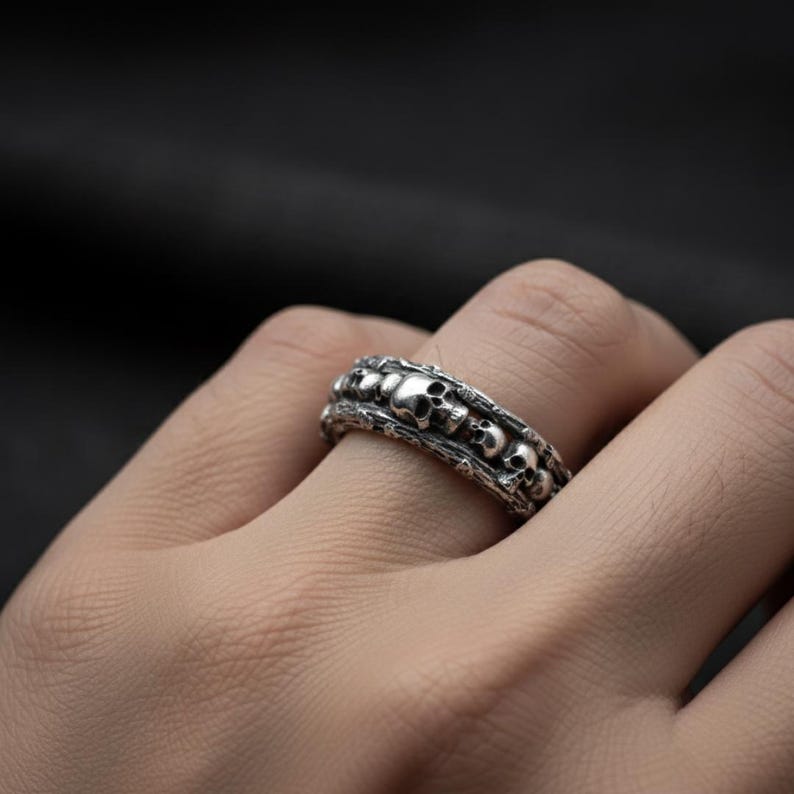 Op de afbeelding: Een zilverkleurige ring met een herhalend schedelontwerp. De ring heeft een getextureerde band en een gedetailleerd, gotisch ontwerp. De schedels zijn klein en gelijkmatig verdeeld over de ring.