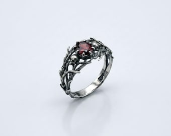 May include: A silver ring with a dark red gemstone at the center. The ring features a detailed design of skulls and foliage. The ring is set against a white background, highlighting its intricate details and gothic style.