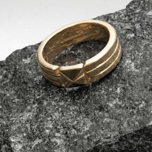 May include: A gold-colored ring with a geometric design. The ring features a series of parallel lines and a central, pointed design. The ring is set against a dark, textured background.