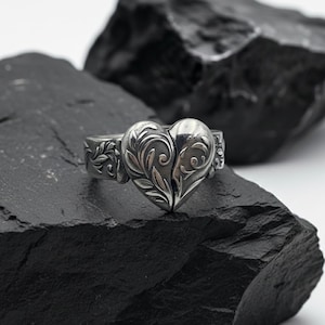 Puede incluir: Un anillo plateado con un diseño de corazón dividido y detalles florales intrincados. El anillo se muestra sobre un fondo oscuro y texturizado, resaltando el brillo del metal y la detallada artesanía de la joya.
