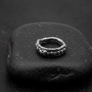 Op de afbeelding: Een zilveren ring met een schedelontwerp. De ring heeft een band met kleine schedels en een getextureerde buitenband. De ring wordt gepresenteerd op een donkere, getextureerde ondergrond, waardoor een contrast ontstaat dat de details benadrukt.