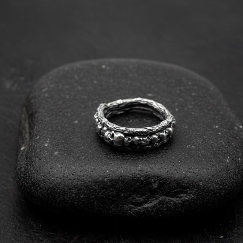 Op de afbeelding: Een zilveren ring met een schedelontwerp. De ring heeft een band met kleine schedels en een getextureerde buitenband. De ring wordt gepresenteerd op een donkere, getextureerde ondergrond, waardoor een contrast ontstaat dat de details benadrukt.