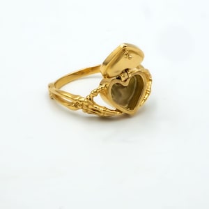 May include: A gold-colored ring with a heart-shaped locket. The ring features a skeleton hand design holding the heart-shaped locket. The locket is open, revealing the interior. The ring is on a white background.