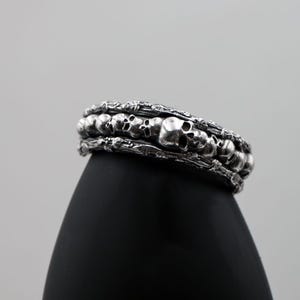 Op de afbeelding: Een zilverkleurige armband met een herhaald schedelontwerp. De armband heeft een gedetailleerd, getextureerd uiterlijk, met meerdere rijen schedels en ingewikkelde details. De schedels zijn gelijkmatig verdeeld en lijken van metaal te zijn gegoten. De armband wordt op een zwart oppervlak weergegeven.