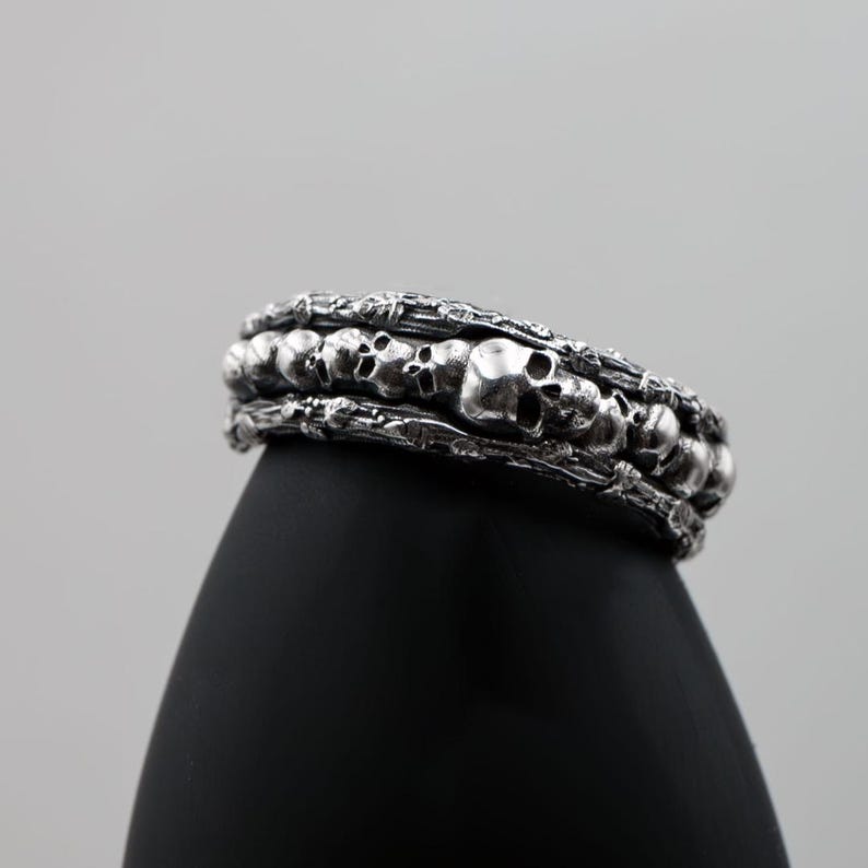 Op de afbeelding: Een zilverkleurige armband met een herhaald schedelontwerp. De armband heeft een gedetailleerd, getextureerd uiterlijk, met meerdere rijen schedels en ingewikkelde details. De schedels zijn gelijkmatig verdeeld en lijken van metaal te zijn gegoten. De armband wordt op een zwart oppervlak weergegeven.