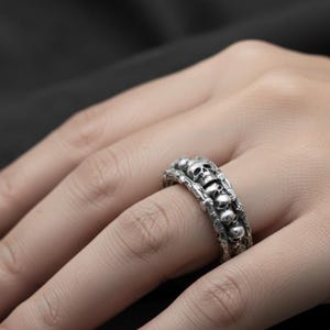 Op de afbeelding: Een zilverkleurige ring met een reeks kleine schedels. De ring heeft een gedetailleerd, gestructureerd ontwerp met de schedels in een rij gerangschikt. De ring wordt aan een vinger gedragen.