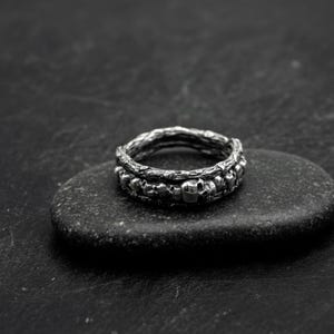 Op de afbeelding: Een zilveren ring met een schedelontwerp. De ring heeft een band van kleine schedels en een getextureerde bovenband. De ring wordt getoond op een donkergrijze steen tegen een zwarte achtergrond.