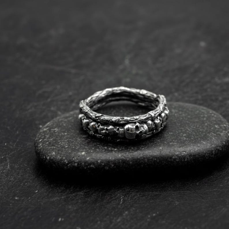 Op de afbeelding: Een zilveren ring met een schedelontwerp. De ring heeft een band van kleine schedels en een getextureerde bovenband. De ring wordt getoond op een donkergrijze steen tegen een zwarte achtergrond.