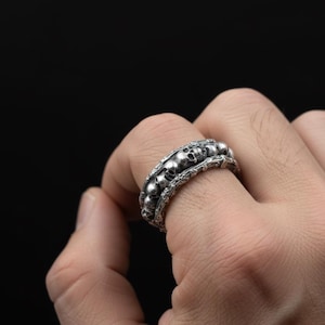 Puede incluir: Un anillo plateado con un diseño de calaveras repetidas. El anillo tiene una banda detallada y texturizada con múltiples calaveras pequeñas que rodean el dedo. El anillo se lleva en una mano sobre un fondo oscuro.
