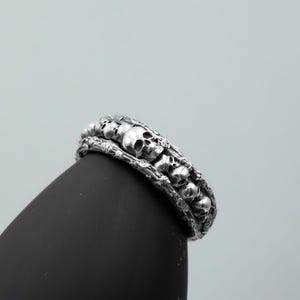 Op de afbeelding: Een zilveren ring met een herhalend schedelontwerp. De ring heeft een gedetailleerd, getextureerd uiterlijk met meerdere kleine schedels die de band omcirkelen. De ring wordt getoond op een zwart oppervlak tegen een lichtgrijze achtergrond.