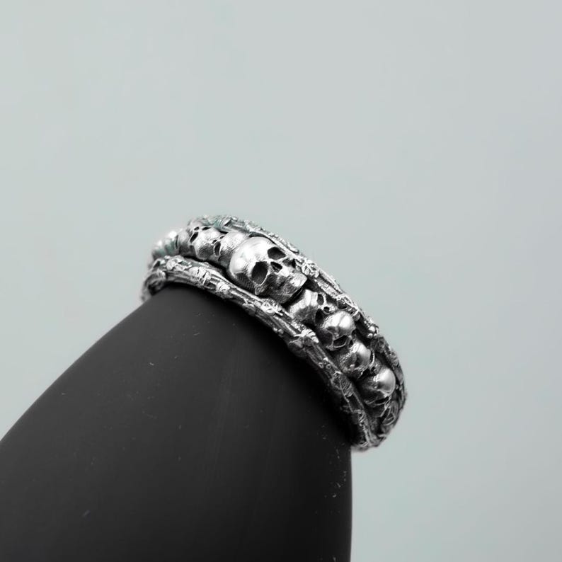 Op de afbeelding: Een zilveren ring met een herhalend schedelontwerp. De ring heeft een gedetailleerd, getextureerd uiterlijk met meerdere kleine schedels die de band omcirkelen. De ring wordt getoond op een zwart oppervlak tegen een lichtgrijze achtergrond.