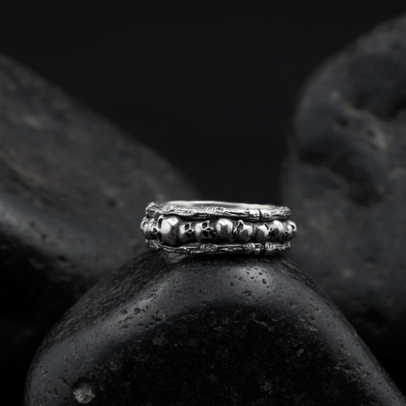 Op de afbeelding: Een zilveren ring met een herhaald schedelontwerp. De ring heeft een gedetailleerd, getextureerd uiterlijk, met meerdere kleine schedels die de band omcirkelen. De ring wordt getoond op een donkere, getextureerde ondergrond.