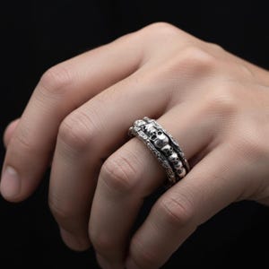 Op de afbeelding: Een zilveren ring met een herhalend schedelontwerp. De ring heeft een getextureerde band en er zijn meerdere schedels zichtbaar. De ring wordt op een vinger gedragen tegen een donkere achtergrond. De ring is een gotische stijl.