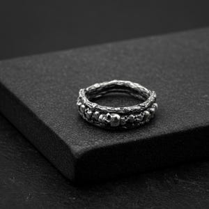 Op de afbeelding: Een zilveren ring met een gedetailleerd ontwerp van kleine schedels en een getextureerde band. De ring heeft een donkere, antieke afwerking en de schedels zijn rond de band gerangschikt. De ring staat tegen een donkere achtergrond.