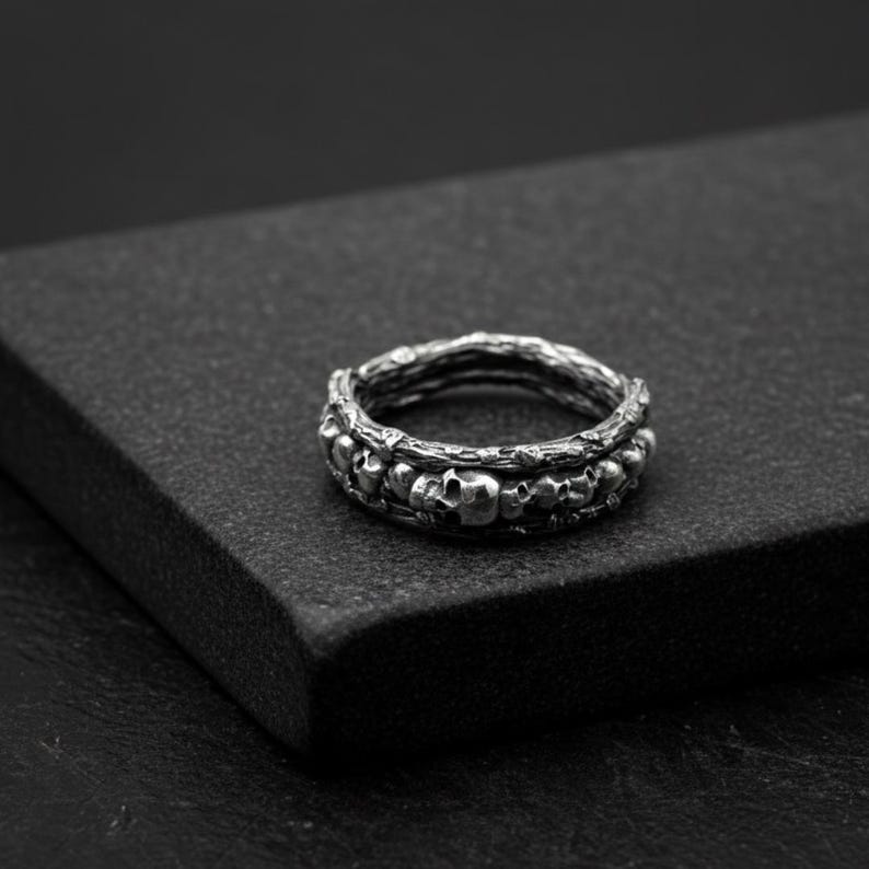 Op de afbeelding: Een zilveren ring met een gedetailleerd ontwerp van kleine schedels en een getextureerde band. De ring heeft een donkere, antieke afwerking en de schedels zijn rond de band gerangschikt. De ring staat tegen een donkere achtergrond.
