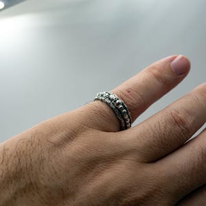 Op de afbeelding: Een zilveren ring met een schedelontwerp wordt aan een vinger gedragen. De ring heeft een getextureerd oppervlak en een gedetailleerd patroon van kleine schedels. De ring is een sieraad, waarschijnlijk voor mode of persoonlijke expressie.