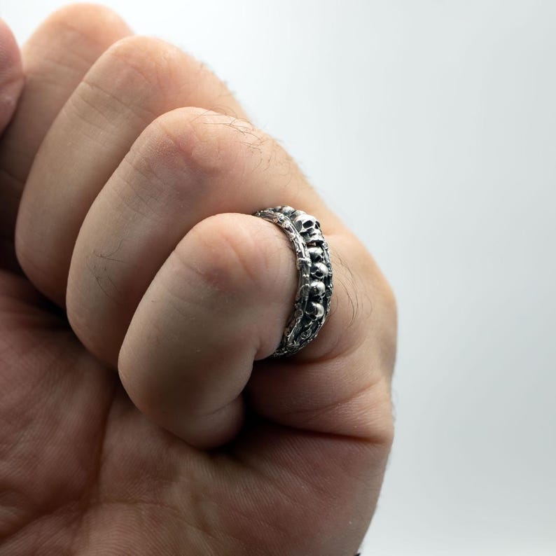 Op de afbeelding: Een zilveren ring met een herhalend schedelpatroon. De ring wordt om een vinger gedragen en toont het gedetailleerde schedelontwerp. De ring heeft een donkere, geoxideerde afwerking die de details verbetert.