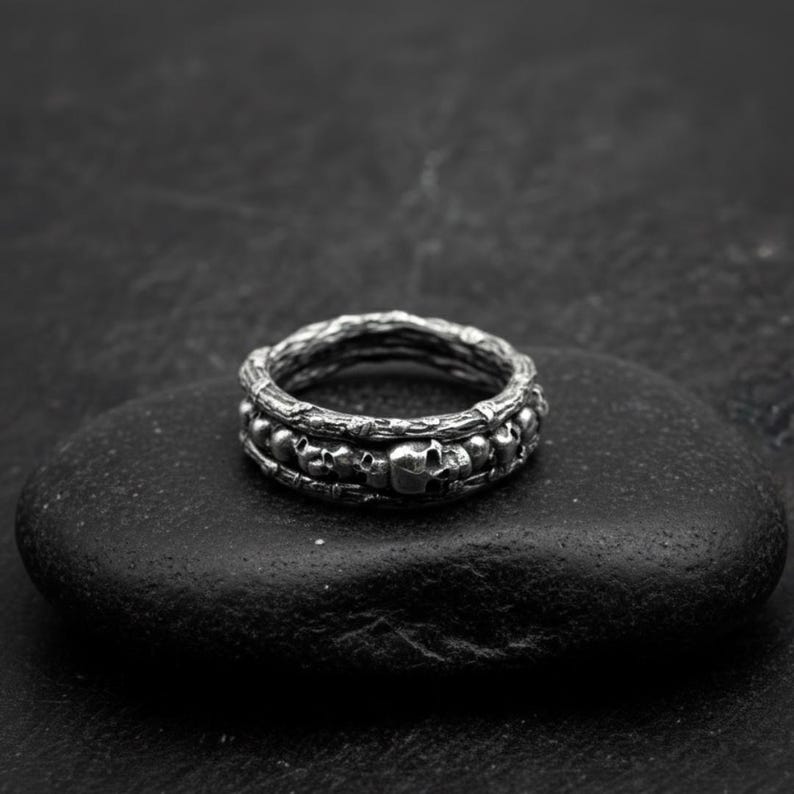 Op de afbeelding: Een zilverkleurige ring met een schedelontwerp. De ring heeft een getextureerde band en een rij kleine schedels rond het midden. De ring wordt gepresenteerd op een donker, getextureerd oppervlak.