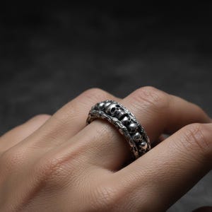 Op de afbeelding: Zilveren ring met een herhaald schedelontwerp. De ring heeft een gedetailleerde, getextureerde band met meerdere kleine schedels die de vinger omsluiten. De ring wordt op een hand gedragen, wat het ingewikkelde vakmanschap en de gotische stijl laat zien.