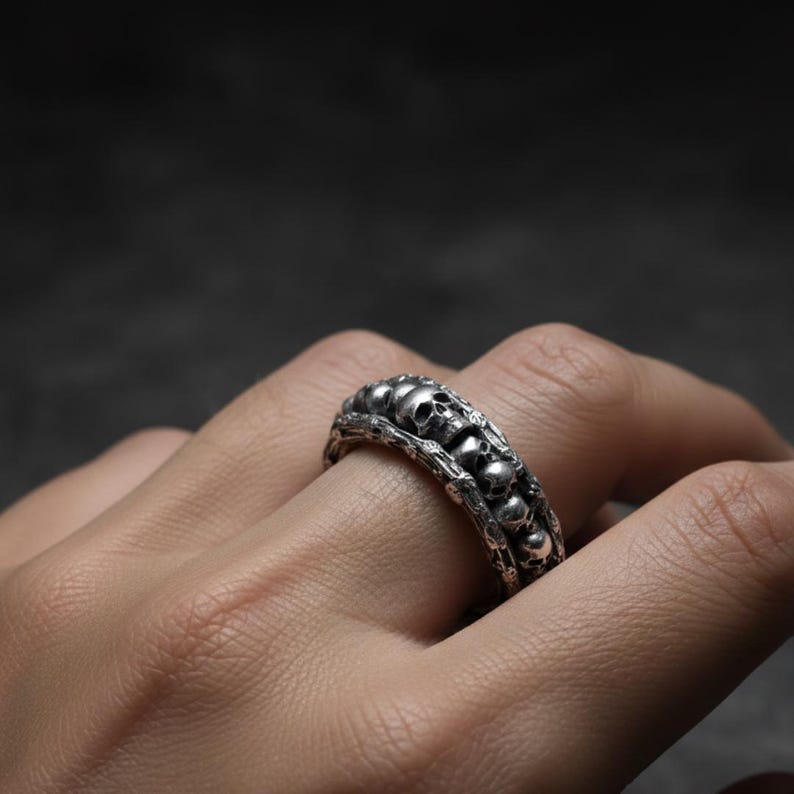 Op de afbeelding: Zilveren ring met een herhaald schedelontwerp. De ring heeft een gedetailleerde, getextureerde band met meerdere kleine schedels die de vinger omsluiten. De ring wordt op een hand gedragen, wat het ingewikkelde vakmanschap en de gotische stijl laat zien.