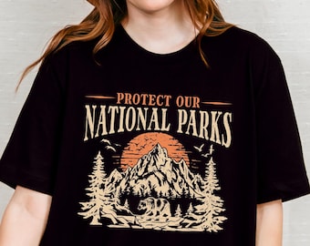 Könnte beinhalten: Schwarzes Oversize-T-Shirt mit einem grafischen Aufdruck einer Bergkette, einem Bären und Bäumen. Der Text "PROTECT OUR NATIONAL PARKS" ist auf dem Shirt aufgedruckt.