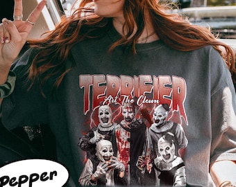 May include: Dark gray t-shirt featuring the text "TERRIFIER Art The Clown" in red and white, with a graphic of several scary clowns. The person in the image is wearing a brown corduroy hat and sunglasses.