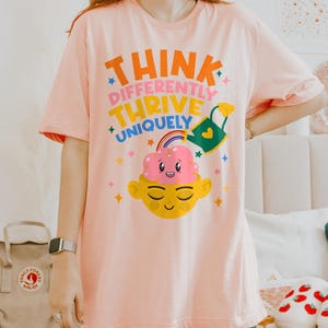 Autism Shirt with a positive message: 'Think Differently, Thrive Uniquely.' This Autism Awareness Shirt features a colorful design with a smiling brain, rainbow, and watering can, symbolizing growth and neurodiversity. Perfect as an Autism Teacher Shirt, Neurodivergent Shirt, ADHD Shirt, Anxiety Shirt, or ABA Therapist Shirt to promote inclusivity and self-expression