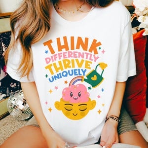 Autism Shirt with a positive message: 'Think Differently, Thrive Uniquely.' This Autism Awareness Shirt features a colorful design with a smiling brain, rainbow, and watering can, symbolizing growth and neurodiversity. Perfect as an Autism Teacher Shirt, Neurodivergent Shirt, ADHD Shirt, Anxiety Shirt, or ABA Therapist Shirt to promote inclusivity and self-expression
