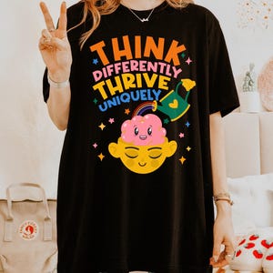 Autism Shirt with a positive message: 'Think Differently, Thrive Uniquely.' This Autism Awareness Shirt features a colorful design with a smiling brain, rainbow, and watering can, symbolizing growth and neurodiversity. Perfect as an Autism Teacher Shirt, Neurodivergent Shirt, ADHD Shirt, Anxiety Shirt, or ABA Therapist Shirt to promote inclusivity and self-expression