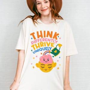 Autism Shirt with a positive message: 'Think Differently, Thrive Uniquely.' This Autism Awareness Shirt features a colorful design with a smiling brain, rainbow, and watering can, symbolizing growth and neurodiversity. Perfect as an Autism Teacher Shirt, Neurodivergent Shirt, ADHD Shirt, Anxiety Shirt, or ABA Therapist Shirt to promote inclusivity and self-expression