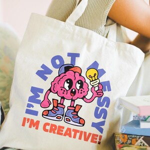 Op de afbeelding: Crèmekleurige draagtas met een cartoonbrein dat een gloeilamp vasthoudt. De tekst op de tas luidt "I'M NOT MESSY I'M CREATIVE!" in blauw en rood. Het brein is roze met blauwe schoenen en een pet.