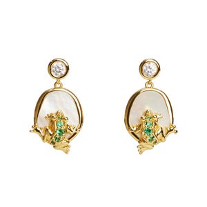 Peut inclure: Paire de boucles d'oreilles dorées avec un motif de grenouille. Chaque boucle d'oreille a une base ovale blanche avec un cadre doré, une petite grenouille verte et une pierre transparente étincelante en haut. La grenouille est ornée de pierres vertes.