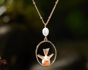 Könnte beinhalten: Eine goldene Halskette mit einer kleinen weißen Perle über einem ovalen goldenen Rahmen. Im Inneren des ovalen Rahmens befindet sich ein Miniatur-Vogel aus Keramik mit weißem Körper, braunen Flügeln und orangefarbener Brust an der Kette.