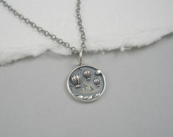 May include: A silver necklace with a round pendant featuring a design of hot air balloons and stars. The pendant is detailed with a textured surface and a small silver bead.