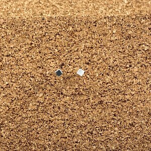 May include: Two small square earrings, one silver and one black, on a brown cork surface.
